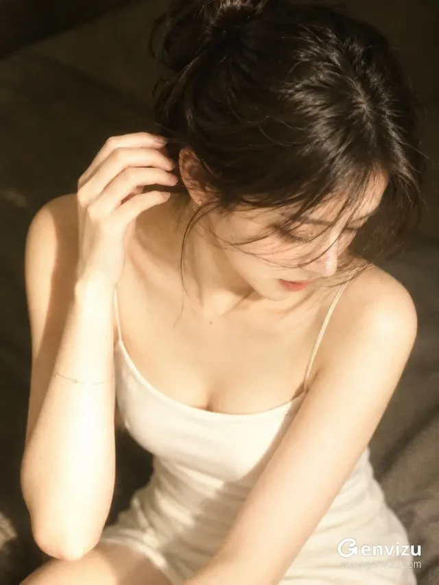 An intimate, sunlit portrait of a young East Asian woman with her head bowed and hair loosely bunned, resting her hand near her ear in a quiet interior.