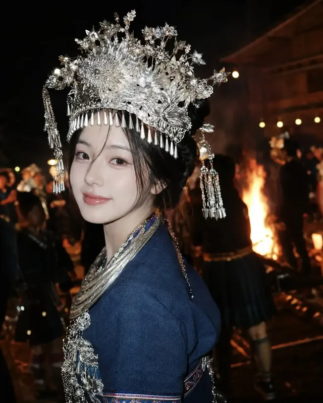 A young Korean woman with dark hair styled up, wearing a large, ornate silver crown with tassels and jewelry, turns to look over her shoulder with a subtle smile. She wears a dark blue ethnic top. A bright bonfire illuminates the background, creating a strong contrast with her silver adornments.
