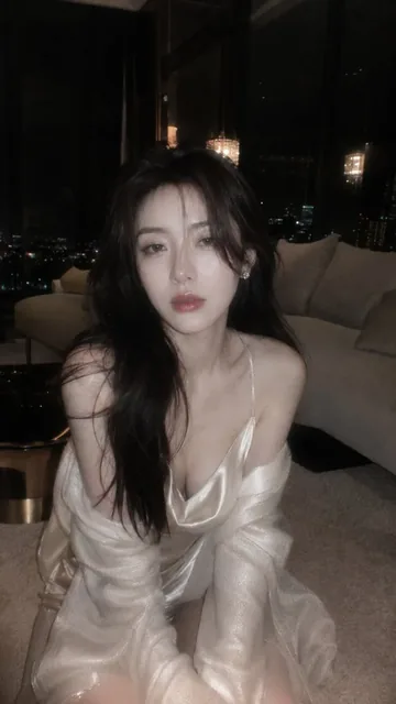 A beautiful young East Asian woman with fair skin and dark hair, wearing satin loungewear, sits on a carpet in a dimly lit luxury penthouse at night, looking calmly at the camera.