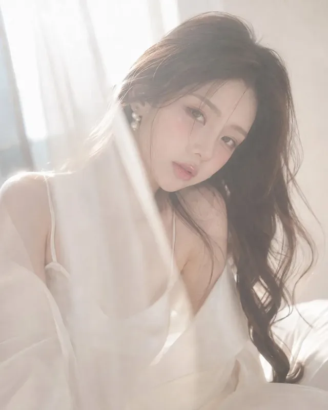 Young Korean female idol with short midface, almond eyes, and full lips, wearing a white satin slip and sheer cover-up, sitting by a window with soft backlight and sheer fabric in the foreground.
