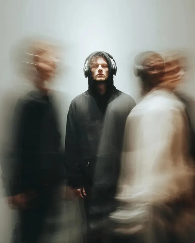 A young man in a black hoodie with headphones stands still in the center of a blurred, fast-moving crowd, staring calmly into the camera.