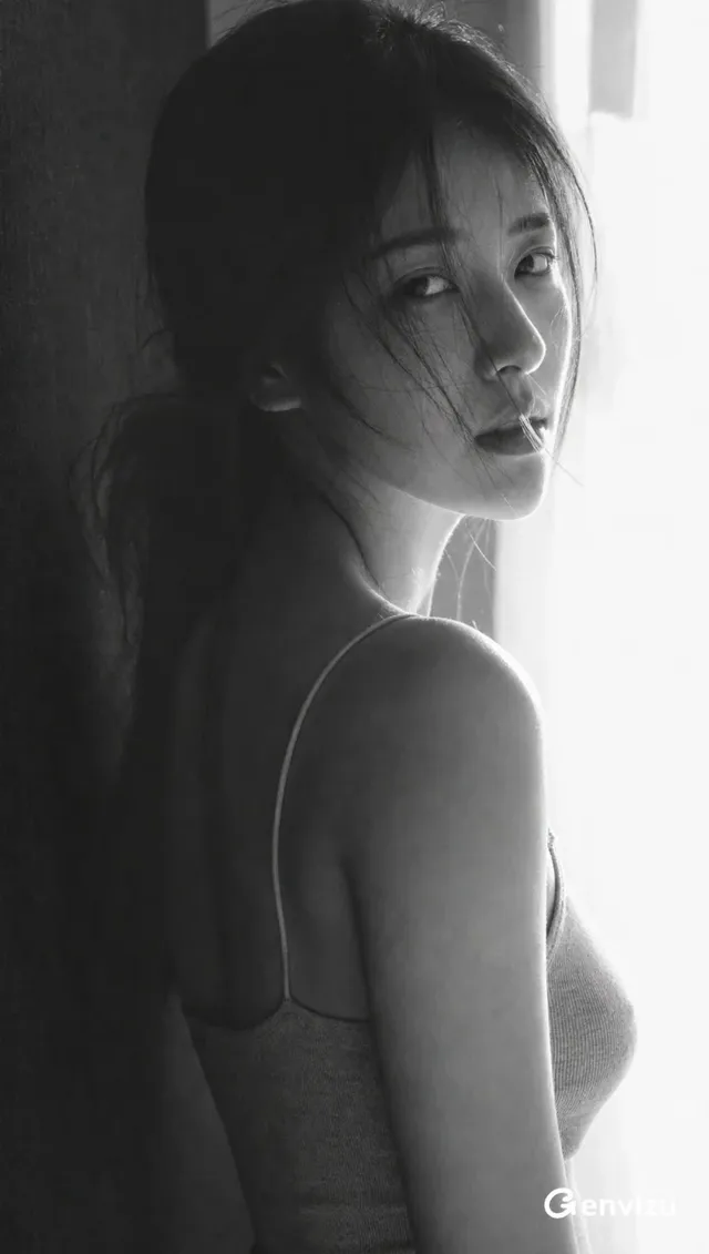 High-contrast black and white portrait of a young East Asian woman, her face turned towards the camera, with stray hairs framing her intense, weary expression. She is illuminated by soft window light from the right.