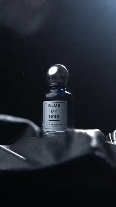 Cinematic commercial for 'BLUE BY ISHQ' luxury perfume. Dark blue bottle with silver cap on leather, mist spray illuminated by spotlight. Features close-ups of label, water drops, finger pressing nozzle, and rotating camera.