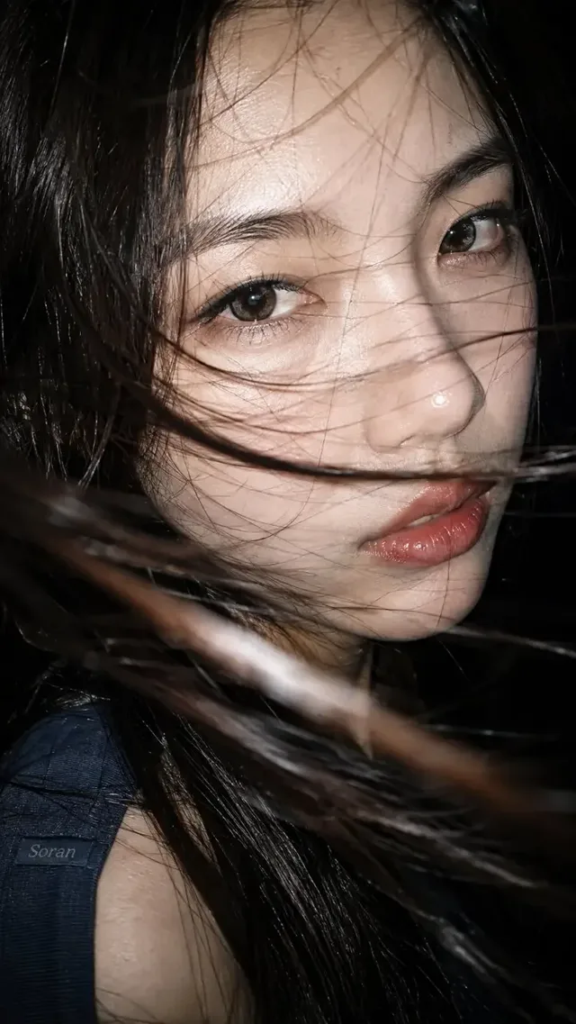Extreme close-up of a young East Asian woman's face, dark hair partially obscuring her features, with sharp specular highlights from a direct flash on her skin and lips, set against a pure black background.
