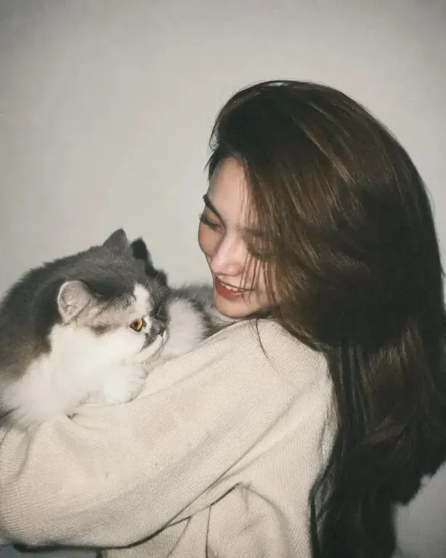 A young Asian woman with long brown hair smiles warmly, holding a white and gray Persian cat. She wears a cream knit sweater. The photo has a vintage, grainy, direct flash style.