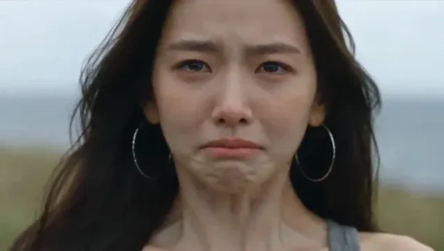 Young East Asian woman in a grey tank top, with large silver hoop earrings, suppresses tears, then pulls her shirt up to cover her mouth and nose, before letting it fall, revealing a weary face.