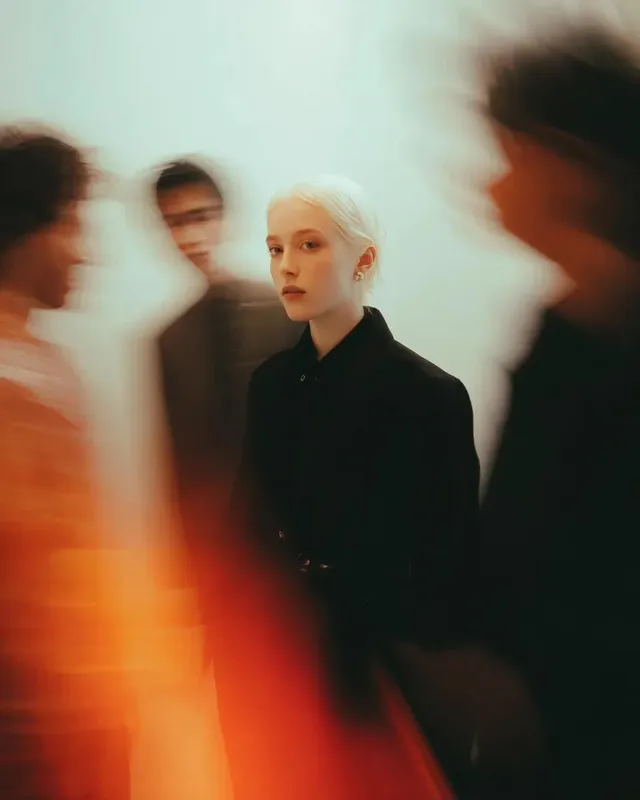 A person stands perfectly still in the center of a minimalist gallery, with blurred figures moving rapidly around them, creating a dramatic motion blur effect. The central figure is sharp and in focus, wearing a black coat and looking directly at the camera.