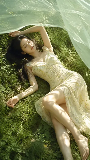 A young woman with closed eyes lies on a grassy field, wearing a light floral dress and a sheer veil, with soft sunlight illuminating the scene.