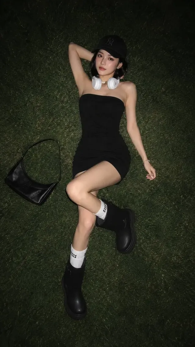 Overhead shot of a young East Asian woman in a black strapless mini dress, baseball cap, and rain boots, lying on green grass at night, illuminated by direct flash with Y2K aesthetic.