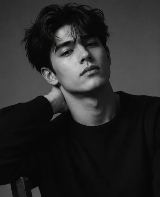 Black and white studio portrait of a young man with dark, voluminous hair, wearing a dark sweater, resting his hand behind his neck, with soft, dramatic lighting.