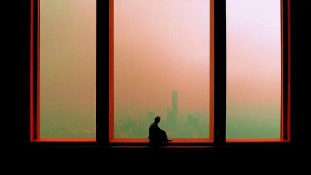 Silhouette of a person sitting on a windowsill looking out at a hazy city skyline.