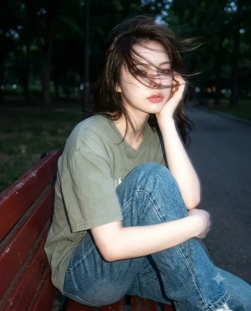 A woman sitting on a park bench, her face mostly obscured by long, dark, windblown hair, in a soft-focus, vintage-style photograph.