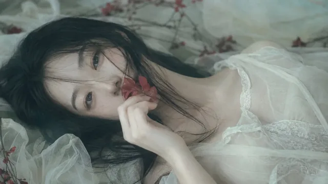 A close-up, slightly overhead shot of a Korean woman with black hair, lying down, her face partially obscured by her hair and a small red flower held to her lips. She has a cold, pale complexion with soft green-blue shadows and creamy highlights, and her eyes gaze softly at the viewer.