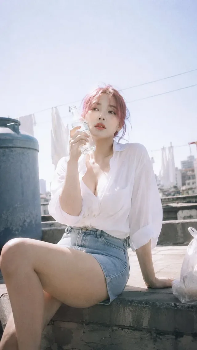 Photorealistic portrait of a young Korean woman with pink hair on a rooftop, wearing an oversized white shirt and denim shorts, winking at the camera. Soft backlight, faded colors, and a 90s Japanese photobook aesthetic.