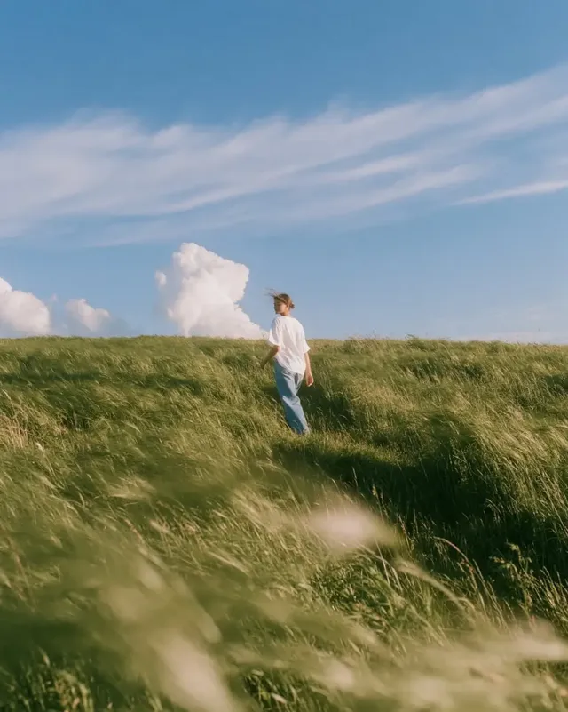 Low angle side view of a young woman in a white tee and jeans walking forward on a path through a vast, swaying green wheat field under a bright blue sky with soft clouds.
