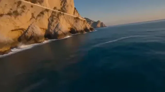 Continuous aerial drone footage of a dramatic coastline with cliffs, a winding road, a village, and sailboats, bathed in golden hour light.