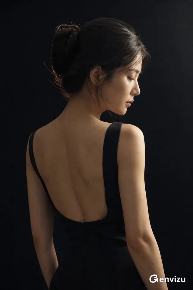 Back view of a 27-year-old East Asian woman in a minimalist black dress, standing or sitting, with her head turned to the side, showcasing her profile and a serene, introspective expression. Soft studio lighting highlights her features and the curve of her back.