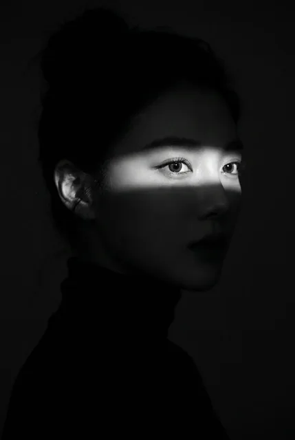 A woman's face in profile, dramatically lit by a sharp horizontal band of light across her eyes and nose, with the rest of her face in deep shadow.