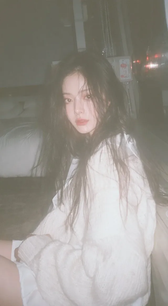 A young Japanese woman with delicate features and long black hair, in casual loungewear, sits by a window, captured in a soft-focus, slightly blurred snapshot with direct flash, evoking a 2000s Japanese photo album feel.
