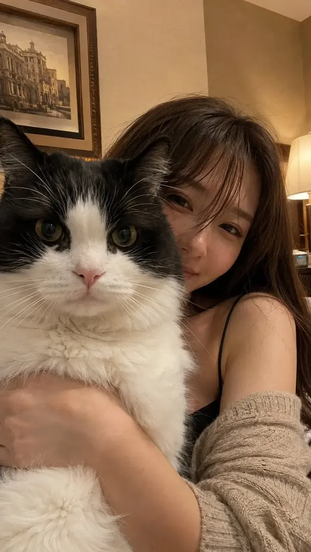 Close-up selfie of a young Japanese woman with long brown hair and a gentle smile, holding a fluffy black and white bicolor cat in front of her face in a dimly lit luxury hotel room.