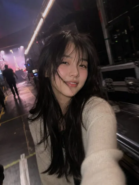 A woman takes a slightly tilted, close-up iPhone selfie in a dimly lit concert backstage hallway, smiling tiredly with flushed cheeks and messy hair, showing motion blur on her arm.