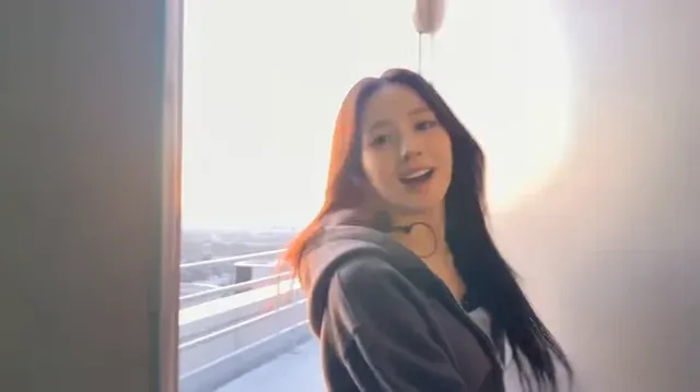 Korean female idol Hana, with long dark hair, smiles at a consumer camcorder on a hidden rooftop, overlooking a city skyline at sunset.