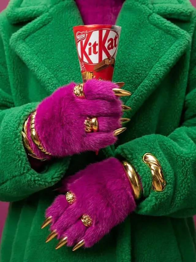 Close-up macro shot of magenta pink furry gloved hands with gold pointed nail tips, wearing an oversized green fleece coat, holding an ice cream cone up close to the camera.