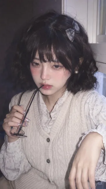 Close-up, soft focus, phone flash portrait of a young woman with short curly hair, wearing glasses and a knitted vest, looking shyly with lips slightly parted around glasses frame.