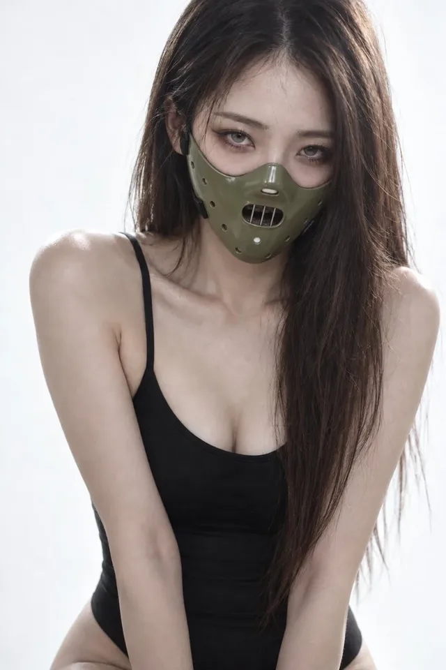 A young woman with dark hair and a metallic mask covering her nose and mouth, wearing a black bodysuit, in a minimalist studio with soft lighting and a hint of red on the mask.