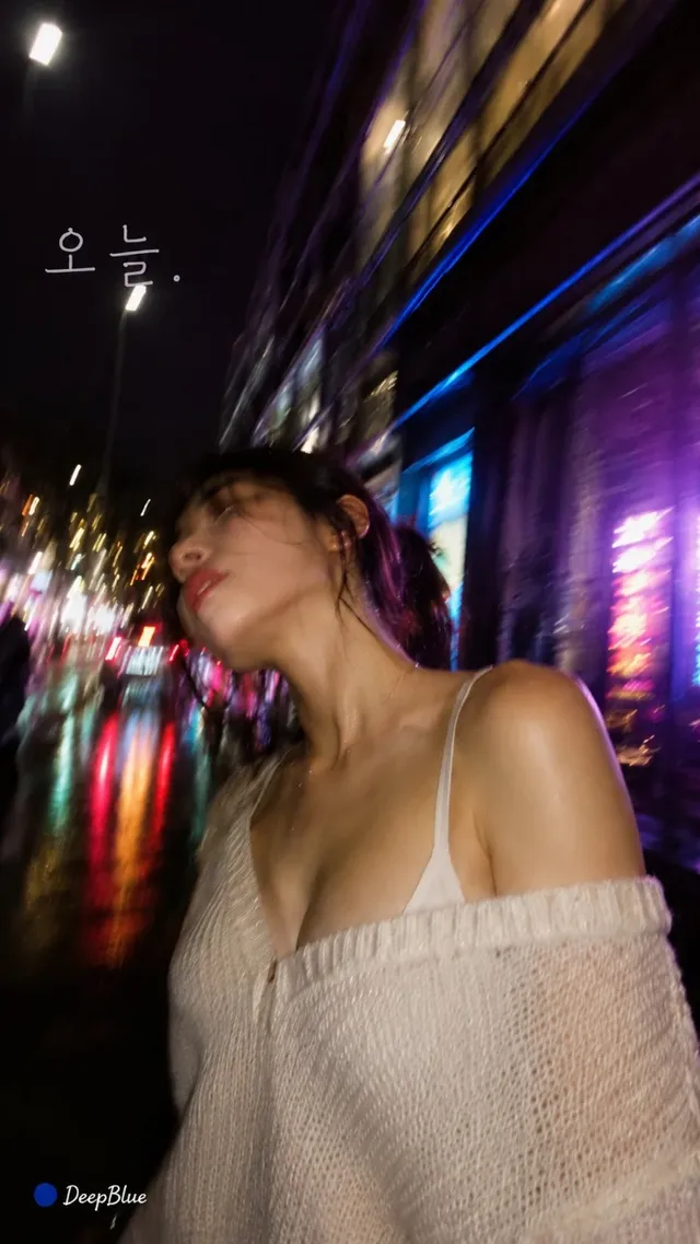 A Korean woman with dark hair in a low bun, wearing a knit top and cardigan, stands on a wet city street at night. Neon lights from buildings and vehicles reflect on the ground, creating a colorful, blurred background. The shot is taken from a low angle, with a slightly tilted perspective.