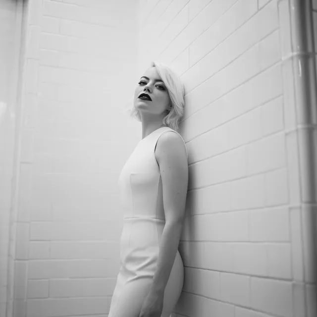 Black and white portrait of Emma Stone with short white-blond hair, black lipstick, wearing a white bodycon dress, against a white tiled wall. Shot on Kodak 35mm film.