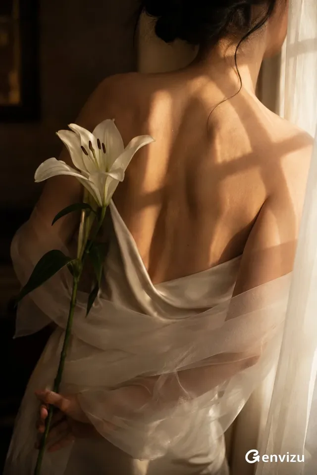 A close-up artistic portrait of a woman's back and shoulders, wearing an ivory silk dress and organza shawl, holding a lily in soft afternoon light.