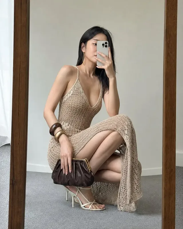 A hyperrealistic 4:5 mirror selfie of a woman in a sand-beige crochet dress, holding a phone and clutch, with an accurate reflection in a wooden-framed mirror.