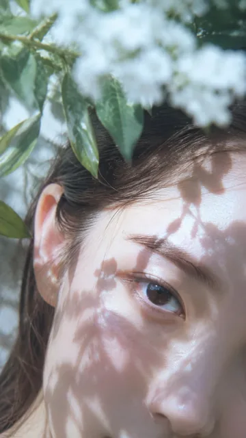 Close-up of a woman's eye and forehead, with dappled light and green leaves in the foreground.