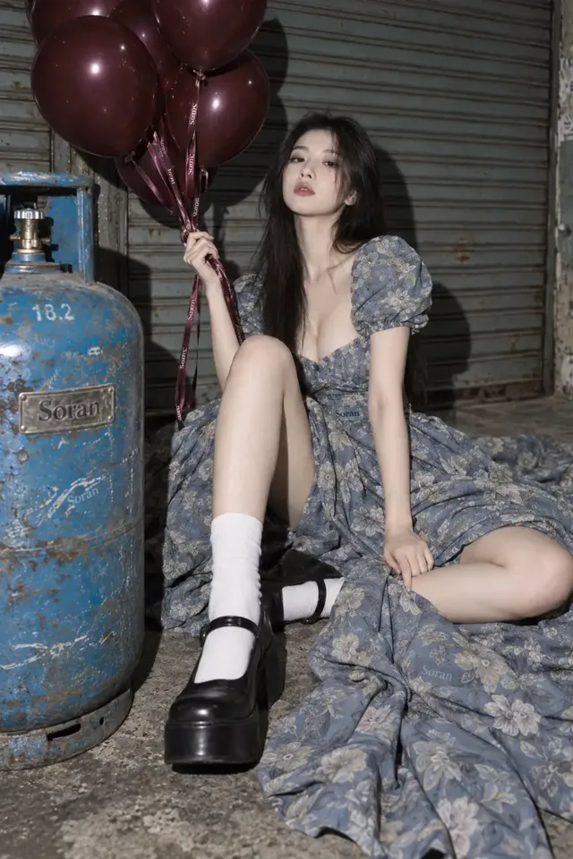 An East Asian woman with long black hair, wearing a large, fog-blue floral print gown with puff sleeves and a cinched waist, sits on the floor of an industrial warehouse. One leg is bent, the other folded, with her arms resting on her knees. She looks directly at the camera with a calm, slightly distant expression. A gas tank is in the foreground, and deep red balloons float to the upper left. The scene has a 90s magazine photoshoot aesthetic with harsh lighting and digital grain.