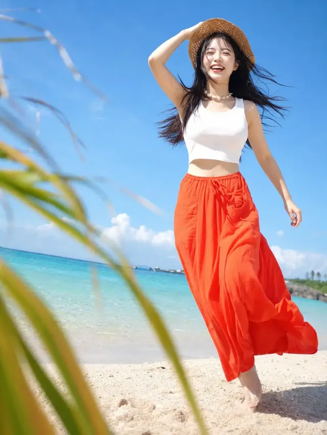 A young Korean woman with dark hair, wearing a white tank top and a flowing orange high-waisted skirt, runs barefoot on a white sandy beach. She is smiling with her mouth open and eyes squinting, one hand holding a straw hat. Sunlight highlights her hair, shoulders, and the edge of her skirt, with a bright blue ocean and sky in the background.