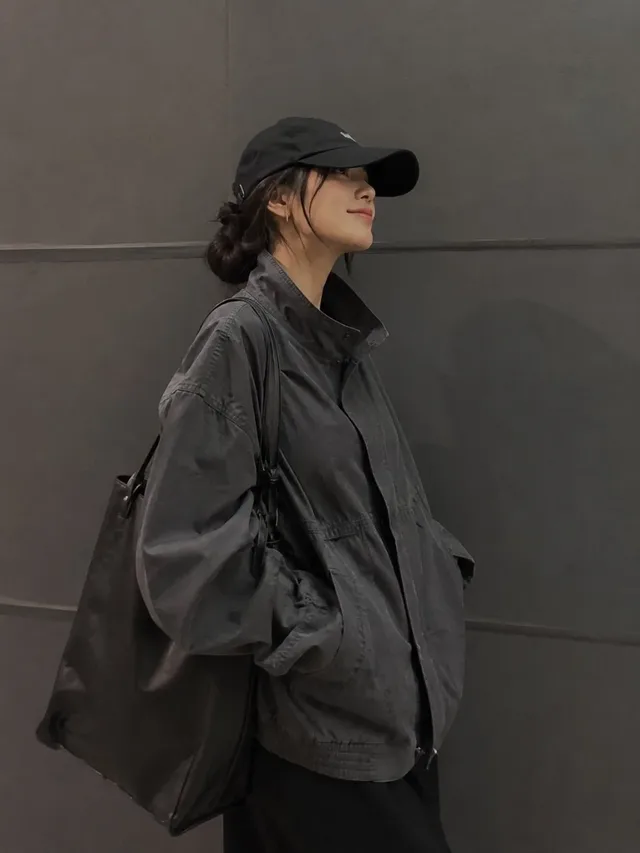 A stylish young woman with a natural smile, wearing a charcoal-gray utility jacket and black baseball cap, stands with hands in pockets against a dark gray wall, hair in a low bun, shallow depth of field.
