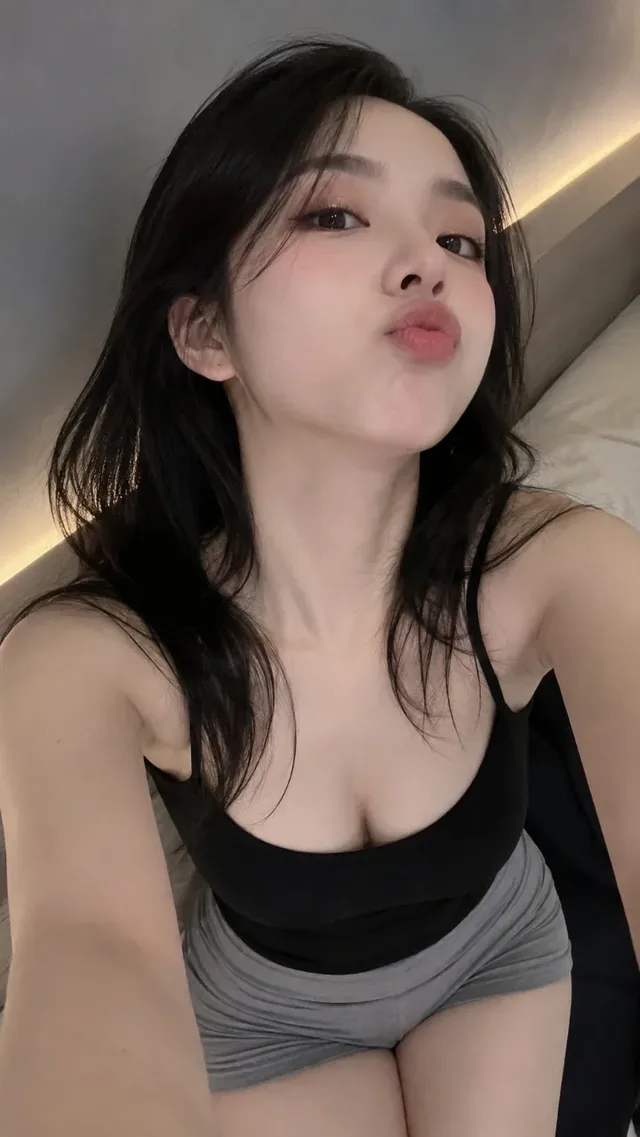 Close-up selfie of a Taiwanese girl with black hair, light blush, and a goofy duck pout, looking up at the camera with ambient lighting.