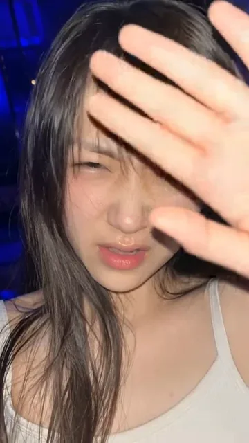 Close-up shot of a young Asian woman reacting to a sudden phone flash, raising her hand to shield her eyes with a mixed expression of annoyance and amusement.