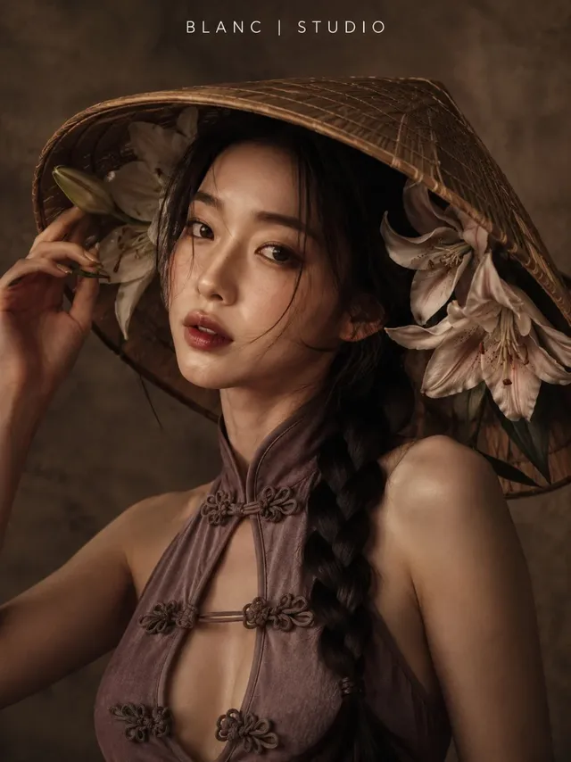 A woman with dark hair in a braid, wearing a mauve purple top and a large conical hat decorated with lilies. She is looking towards the camera with a soft, warm studio light.
