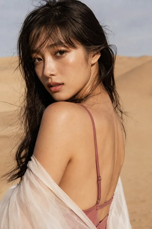 Close-up portrait of a young Japanese woman in a dusty rose swimsuit and cream chiffon shawl, looking back over her shoulder in harsh desert sunlight with blurred dunes in the background.