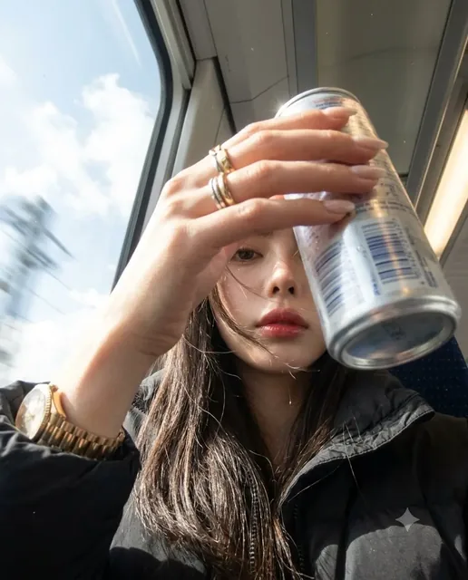 A motion-blurred, early 2000s-style selfie taken inside a moving train. A hand holding a silver energy drink can partially obscures the face, revealing only glimpses of an eye and