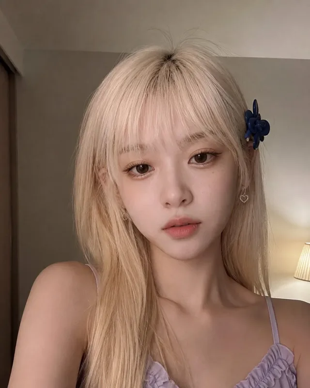 Close-up realistic smartphone selfie of a young woman with blonde hair in a bedroom, looking directly at the camera with a quiet expression. Natural indoor lighting highlights her features and skin texture.
