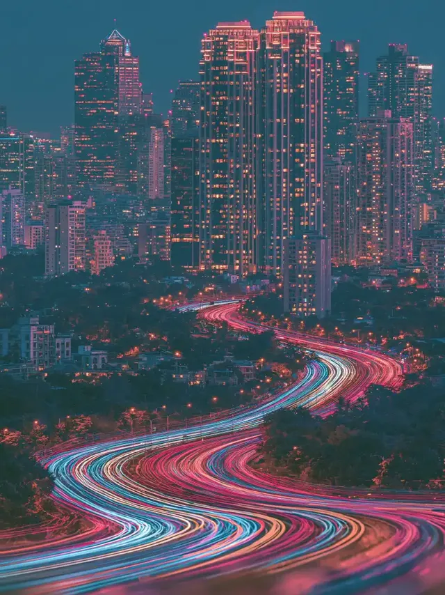 A nighttime cityscape with illuminated skyscrapers and a highway displaying vibrant, colorful light trails from traffic.