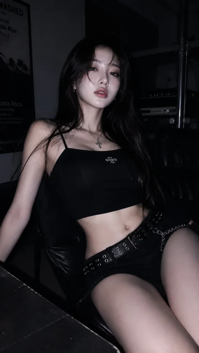 A young woman with dark hair and striking makeup, wearing a black crop top and shorts, poses in a dimly lit room with a dark, edgy aesthetic.