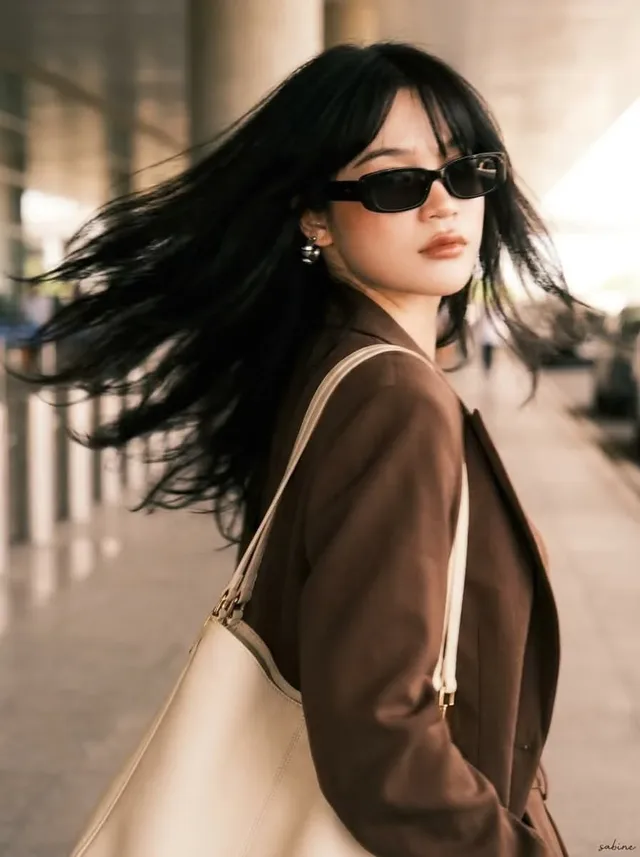 Stylish young woman with long black hair and curtain bangs, wearing a chocolate-brown oversized blazer and matching trousers, holding an ivory tote bag, standing in an open-air parking structure with blurred cars.