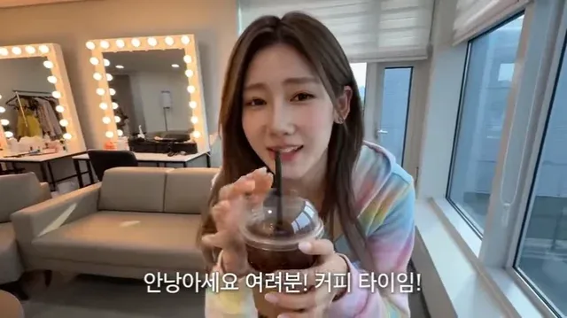 A smiling Korean female idol in a pastel outfit sits in a cozy dressing room, holding an iced coffee and her phone, decorating her cup with a sticker.