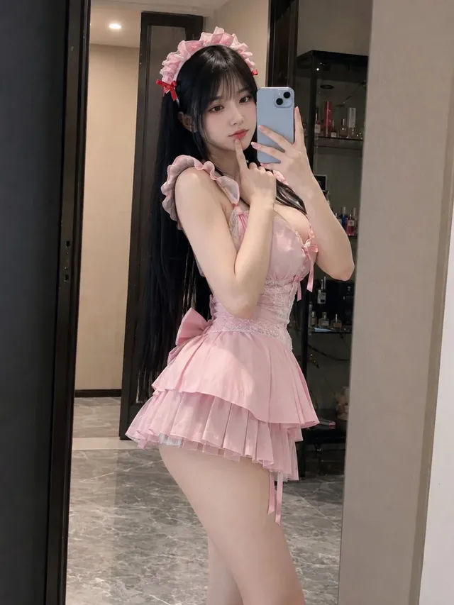 Photorealistic mirror selfie of a beautiful East Asian woman with long black hair, wearing a pale-pink maid dress and headband, posing shyly with a smartphone in a modern apartment.