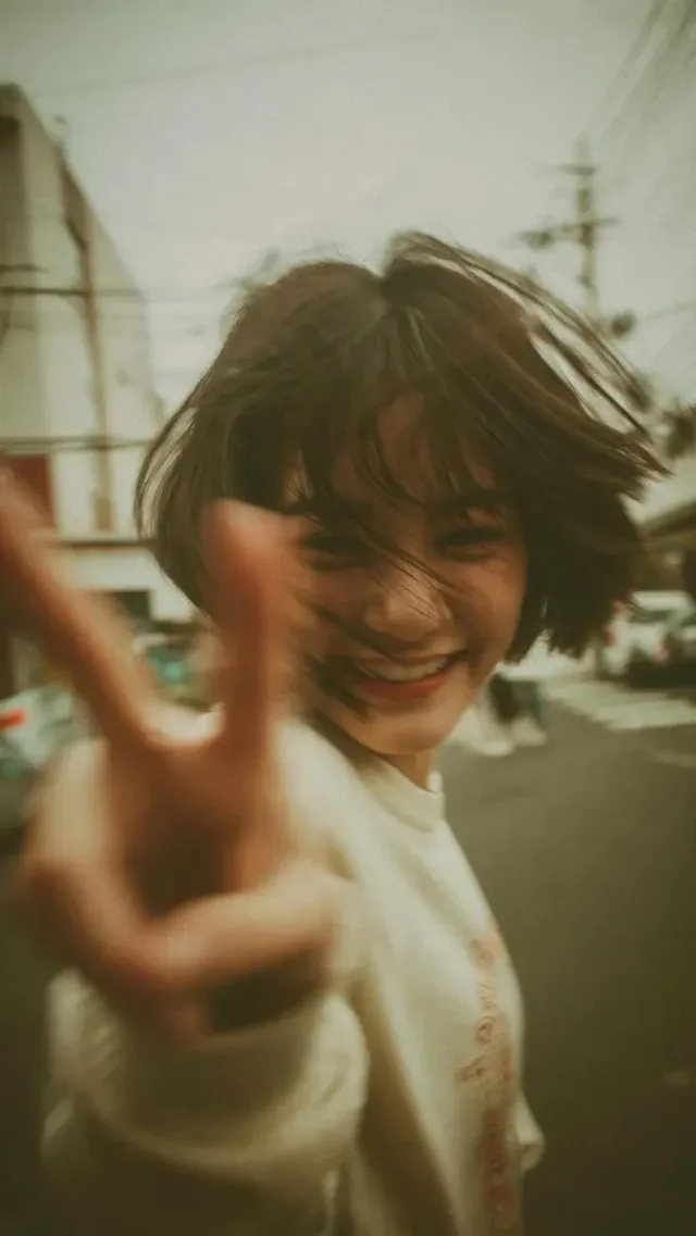 A young woman with a messy bob haircut laughs and flashes a peace sign directly at the camera, her hand in the foreground, on a Japanese street with Y2K aesthetic and film grain.