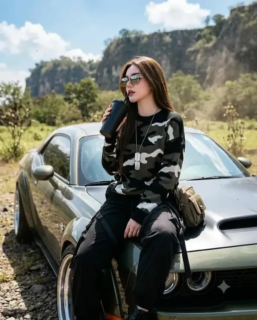 A young woman with long black hair and sunglasses leans against a modified Dodge Challenger, wearing a camouflage sweater and tactical pants, with a scenic rocky cliff background.